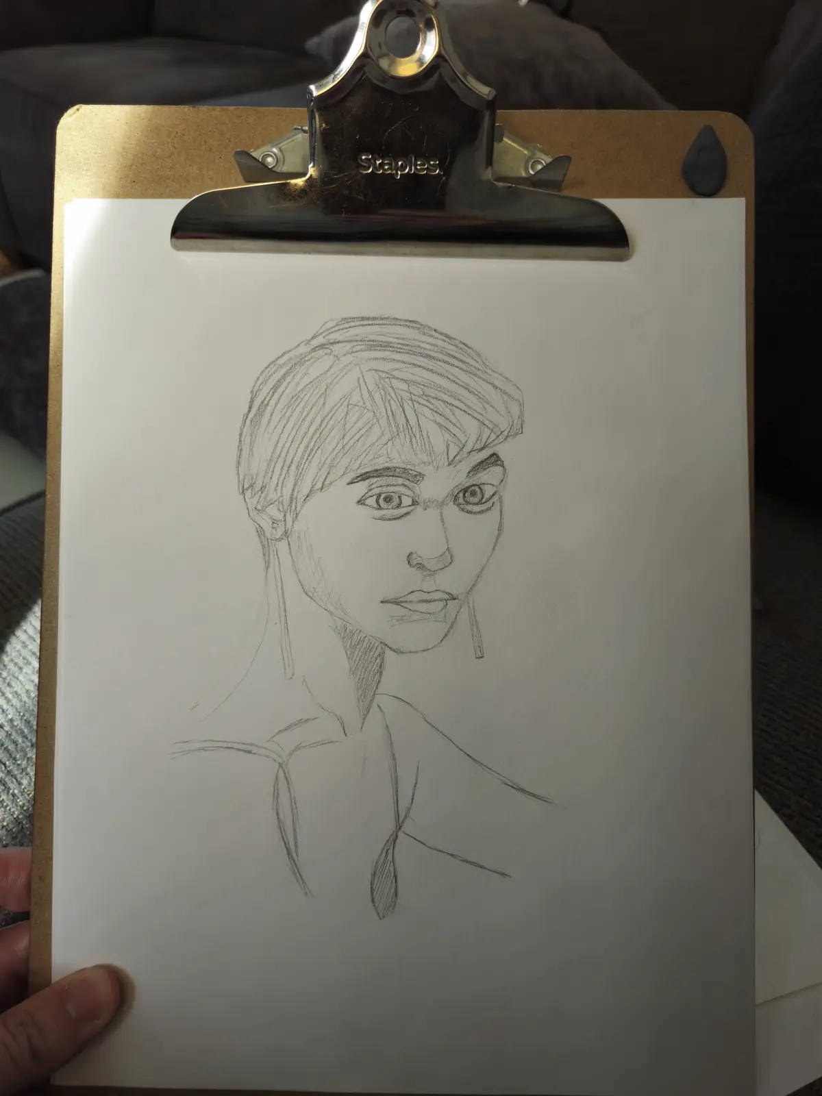 A hand holds a Staples clipboard displaying a pencil sketch of a person's head and shoulders. The drawing shows short hair…