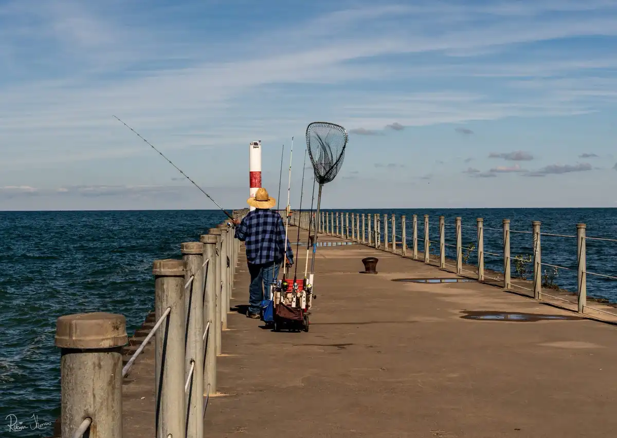 A person in a straw hat and blue plaid shirt walks away from the camera down a concrete pier, holding a fishing rod while…