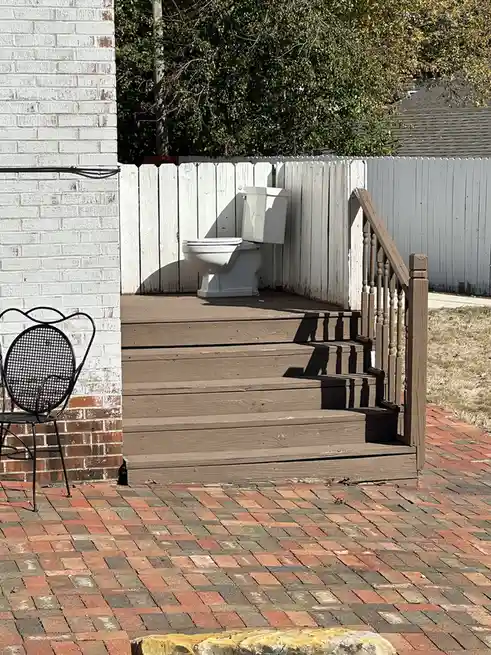 A white toilet sits on a wooden deck behind a white fence, with stairs leading up to it and a brick patio in the foreground…