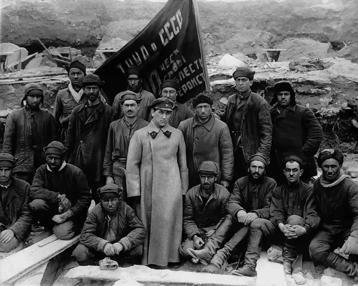 A black-and-white historical photo shows a group of men in work clothes posing with a large flag that reads 'Труд в СССР'…