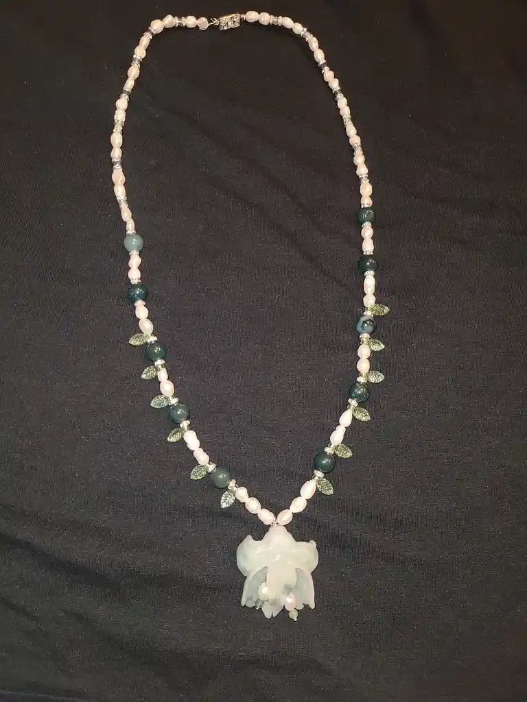 A handmade necklace with alternating pink pearls and green beads, accented with small leaf charms, rests on a black fabric…