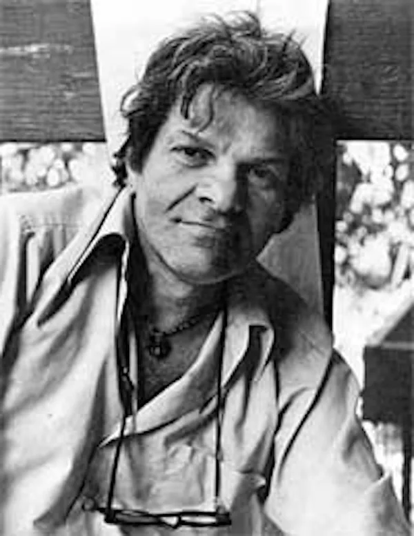 A black-and-white photo shows a man with tousled hair, wearing an open shirt and glasses hanging from his neck, looking…