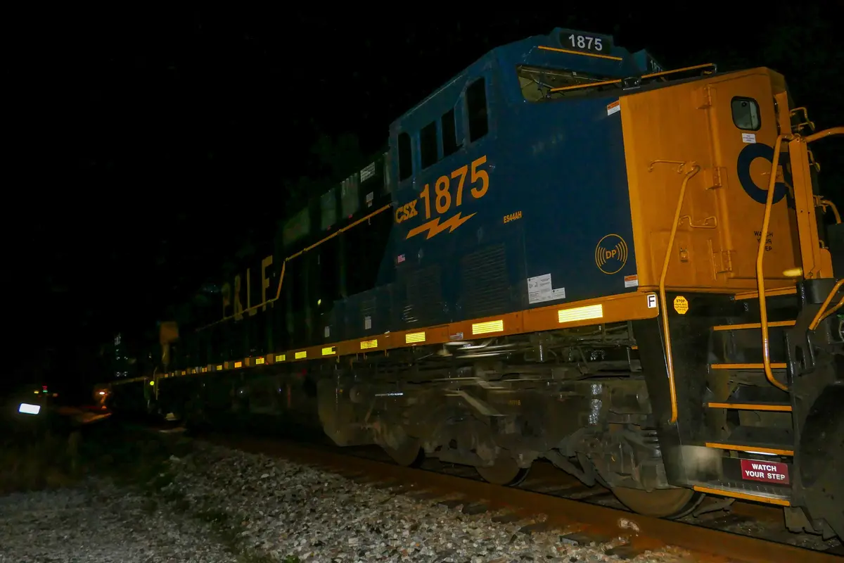 A nighttime photo of a CSX freight train locomotive, number 1875, on tracks with gravel ballast. The train is blue and…