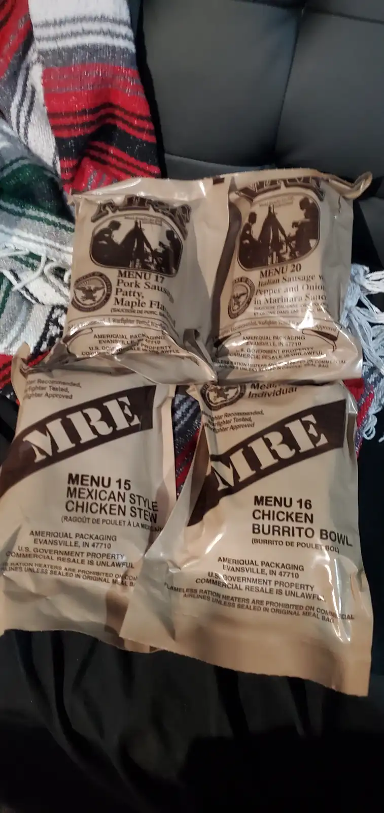 Four U.S. military MRE (Meal, Ready-to-Eat) ration packs are arranged on a patterned blanket, showing menus 15, 16, 17, and…
