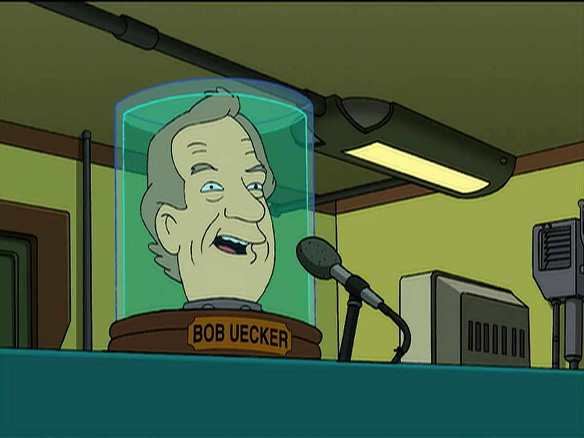 A cartoon image shows a preserved head of Bob Uecker encased in a glass dome on a podium, with a microphone positioned in…