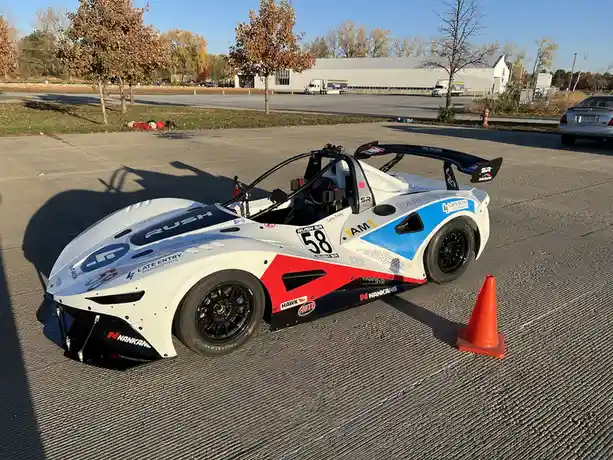A white, red, and blue race car with the number 58 parked on a textured asphalt surface next to an orange traffic cone. The…