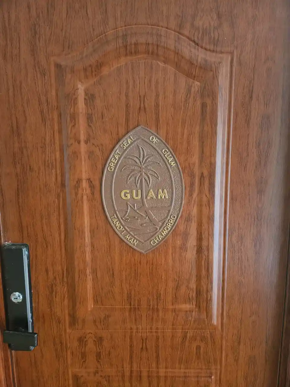 A close-up of a wooden door featuring the Great Seal of Guam, which includes a palm tree, a sailboat, and the text 'GREAT…