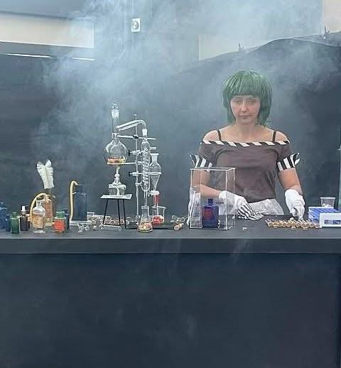 A person with a green bob wig and striped shirt stands behind a lab bench filled with glassware, beakers, and smoking…