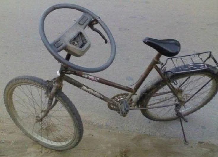 A rusty, old bicycle has a car steering wheel bolted to its handlebar area, replacing the normal handlebars. The bike is on…