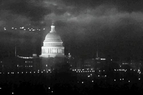 a black and white image of the u.s. capitol building at night, illuminated against a dark sky with clouds. a flock of birds…
