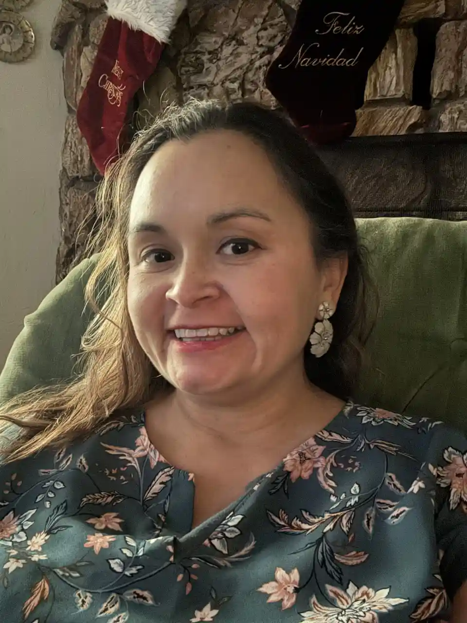 A woman with long brown hair and floral top smiles at the camera while seated in a green chair, with two Christmas stockings…