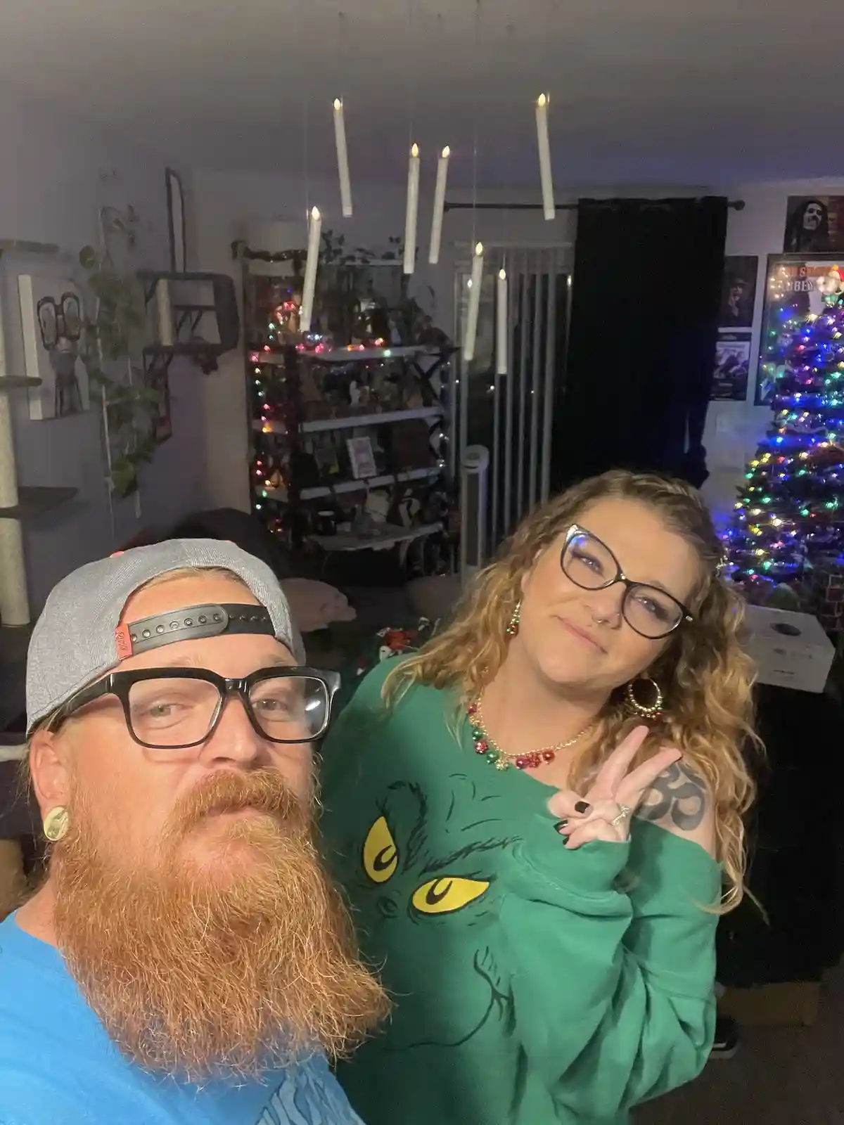 A man with a red beard and glasses wearing a grey cap takes a selfie with a woman in a green Grinch-themed sweater. She…