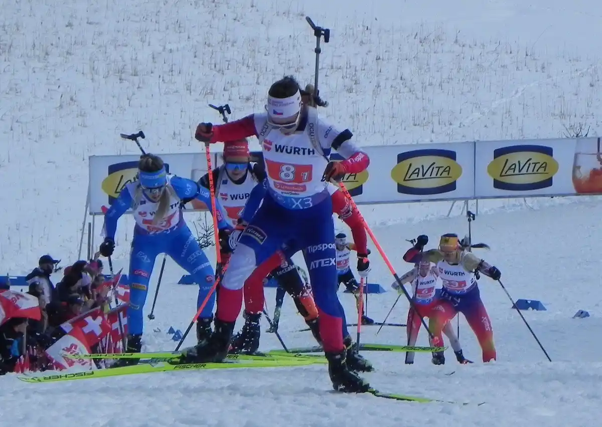 A biathlete in a blue and red uniform with bib number 81 is mid-stride in a snowy race, surrounded by other competitors. The…