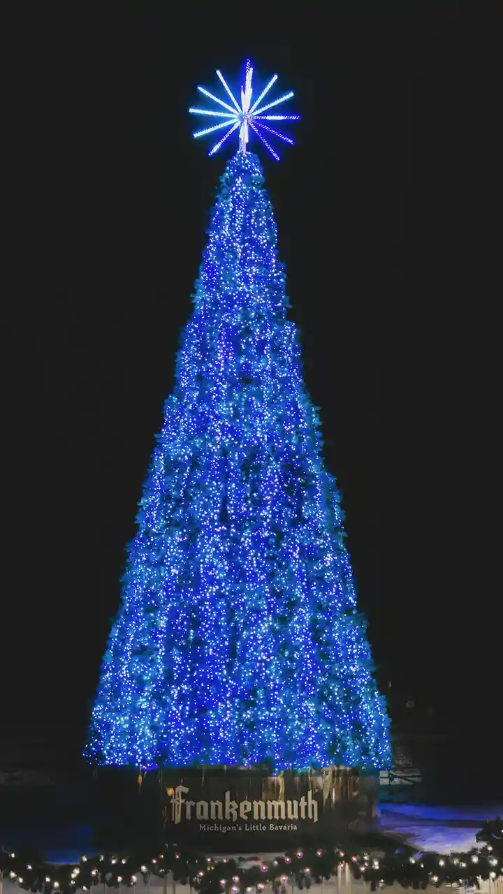 A tall, conical Christmas tree covered in bright blue lights stands against a dark night sky, topped with a star-shaped…