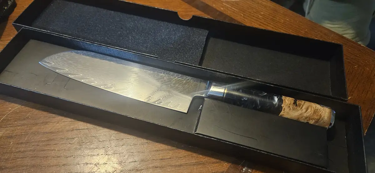 A high-end chef's knife with a damascus steel blade and wooden handle rests in its black presentation box on a wooden table…