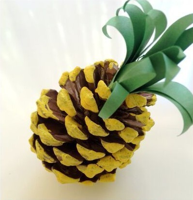A pinecone has been painted yellow and brown to resemble a pineapple, with green paper leaves glued on top. The image is…