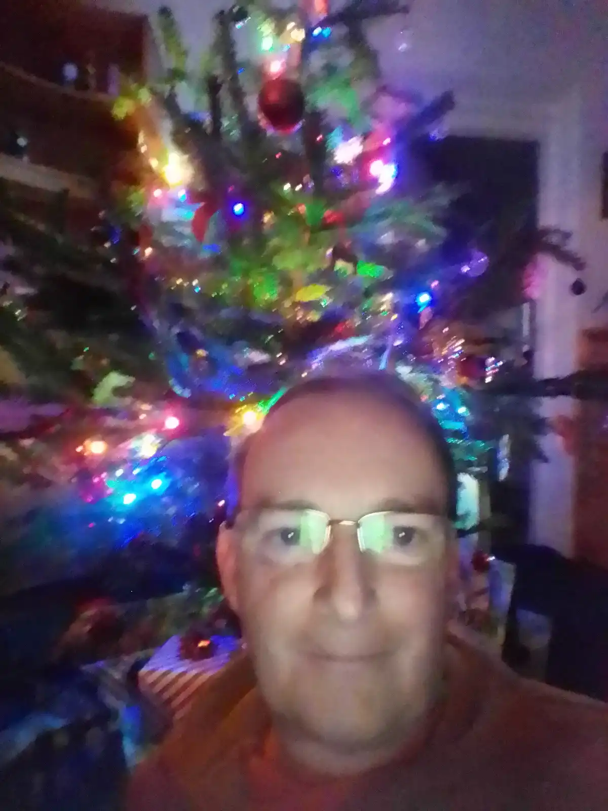 A blurry selfie of a man with glasses smiling at the camera, positioned in front of a brightly lit, decorated christmas tree…