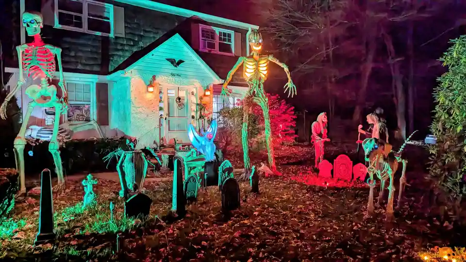 A house at night is covered in elaborate, glowing Halloween decorations including large skeletons, tombstones, a glowing…