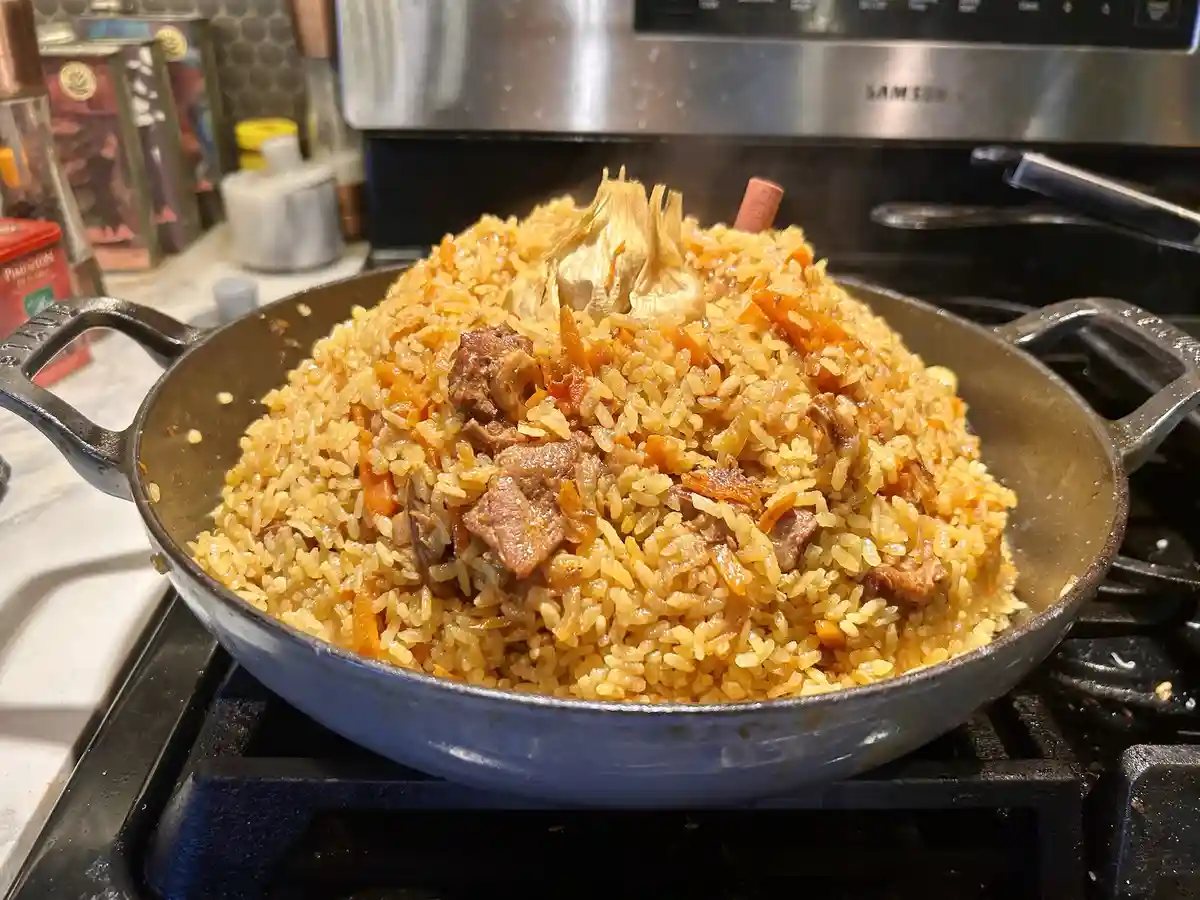 A close-up photo shows a large, steaming cast-iron skillet on a stovetop, filled with a mound of golden-brown rice mixed…