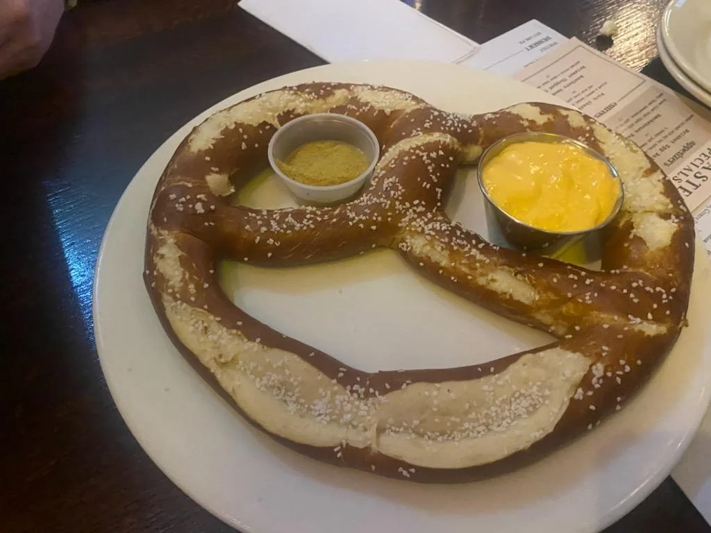 A large, salted pretzel on a white plate has two small cups of dipping sauce—one with yellow mustard and one with orange…