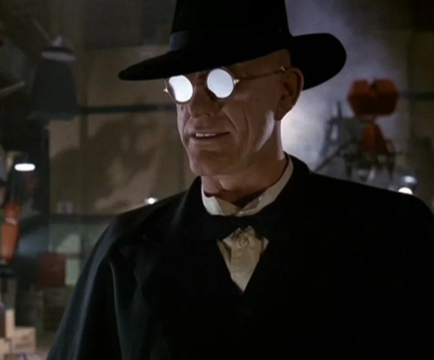 a bald man in a black fedora and dark suit wears round, reflective glasses that glow white, smiling slightly while standing…