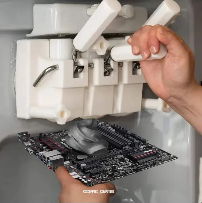A hand holds a computer motherboard directly under a white plastic toilet flush valve mechanism, with another hand gripping…