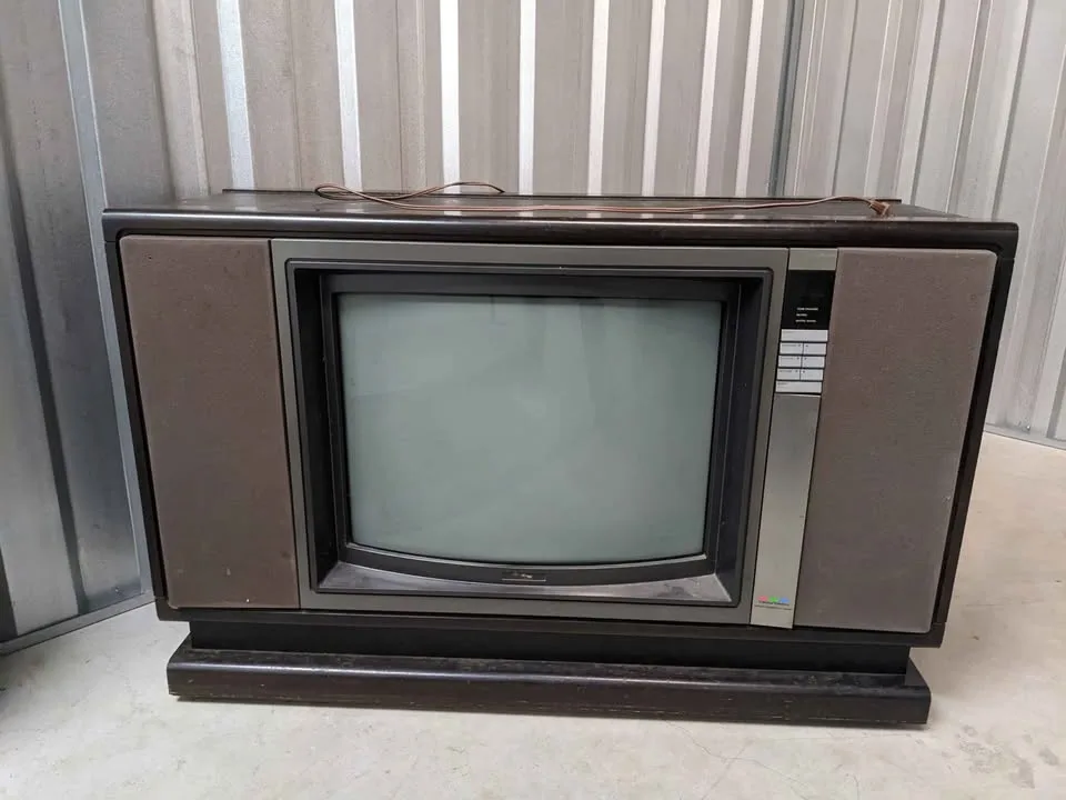 An old, bulky CRT television with a dark screen sits on a stand against a corrugated metal wall. A thin wire lies across the…