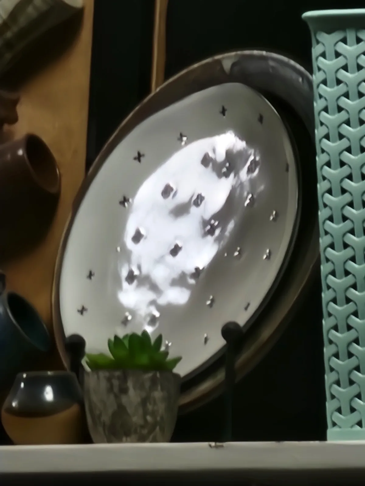 A white ceramic plate with a dark rim and small star-shaped cutouts is displayed on a shelf, reflecting a bright, cloudy…