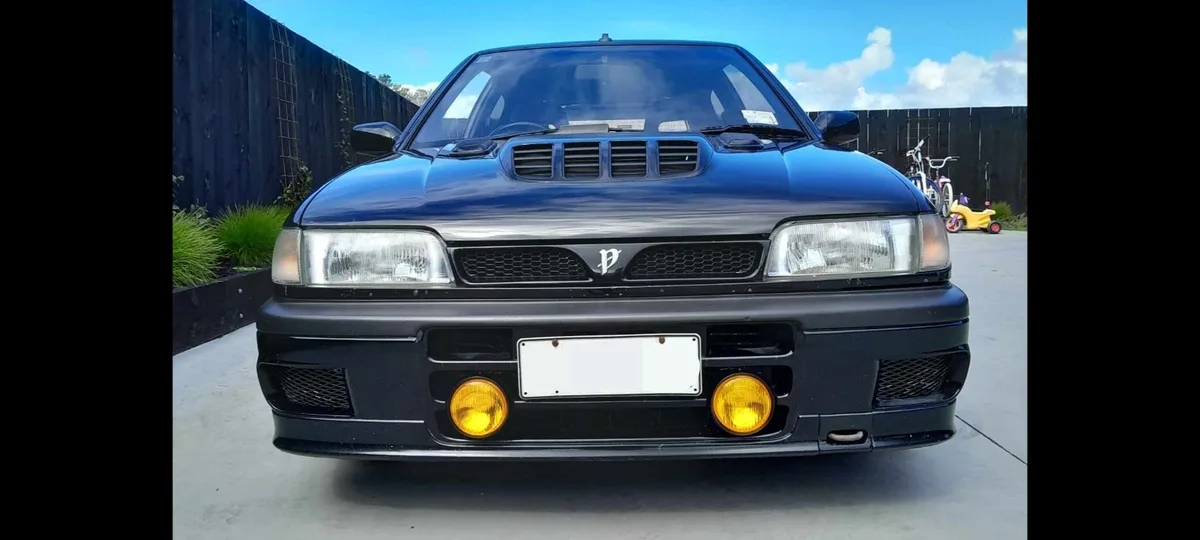 a front-facing photo of a dark blue car with a hood scoop, round yellow fog lights, and a grille with a 'P' logo, parked…