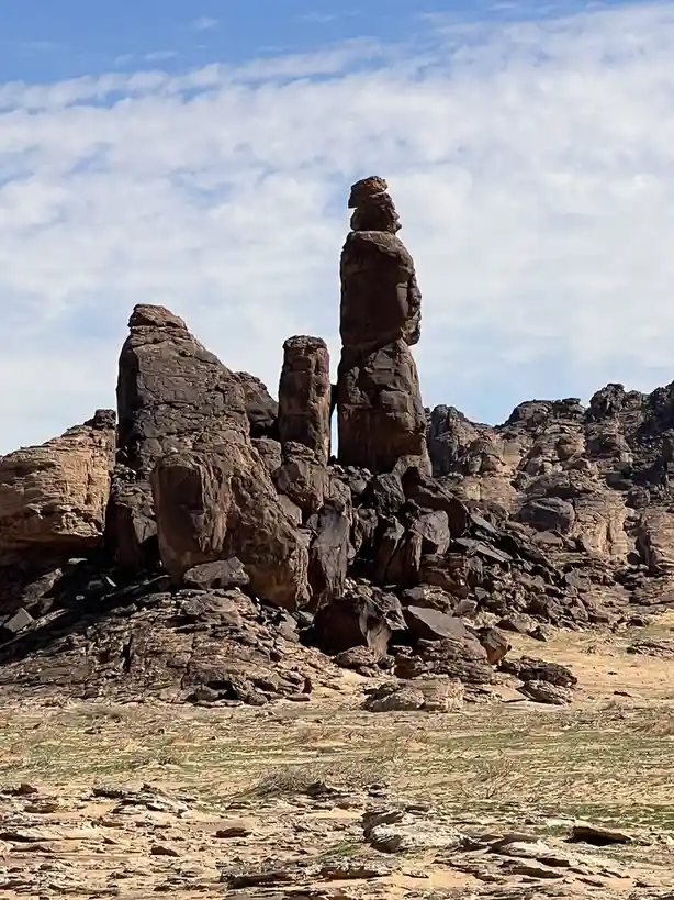 A desert landscape features a tall, slender rock formation that visually resembles a human figure standing upright…