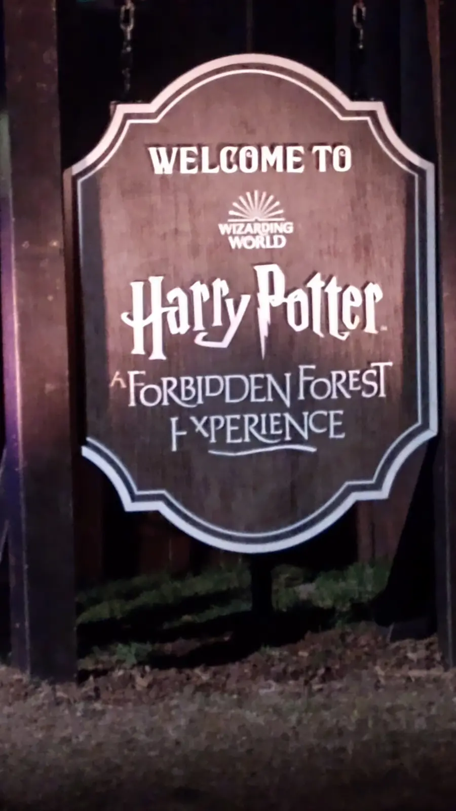 A wooden sign with white lettering reads 'WELCOME TO WIZARDING WORLD Harry Potter A FORBIDDEN FOREST XPERIENCE', where the…