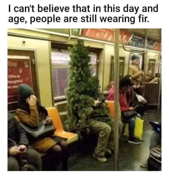 A person sitting on a subway train is completely covered in a green fir tree, with only their legs and feet visible beneath…