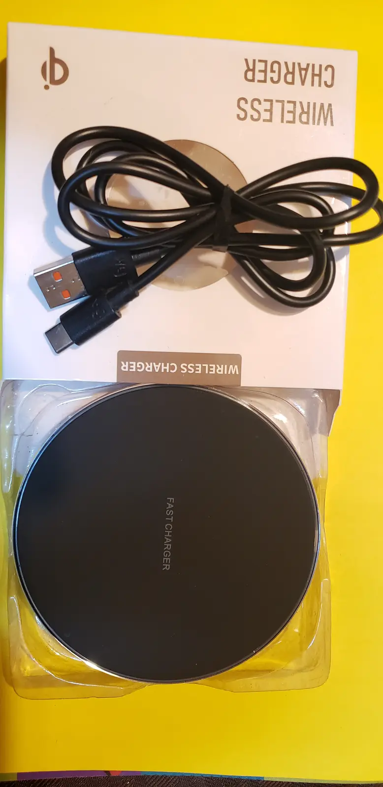 A circular black wireless charger in its packaging, with a USB cable included, has the text 'FAST CHARGER' printed on its…