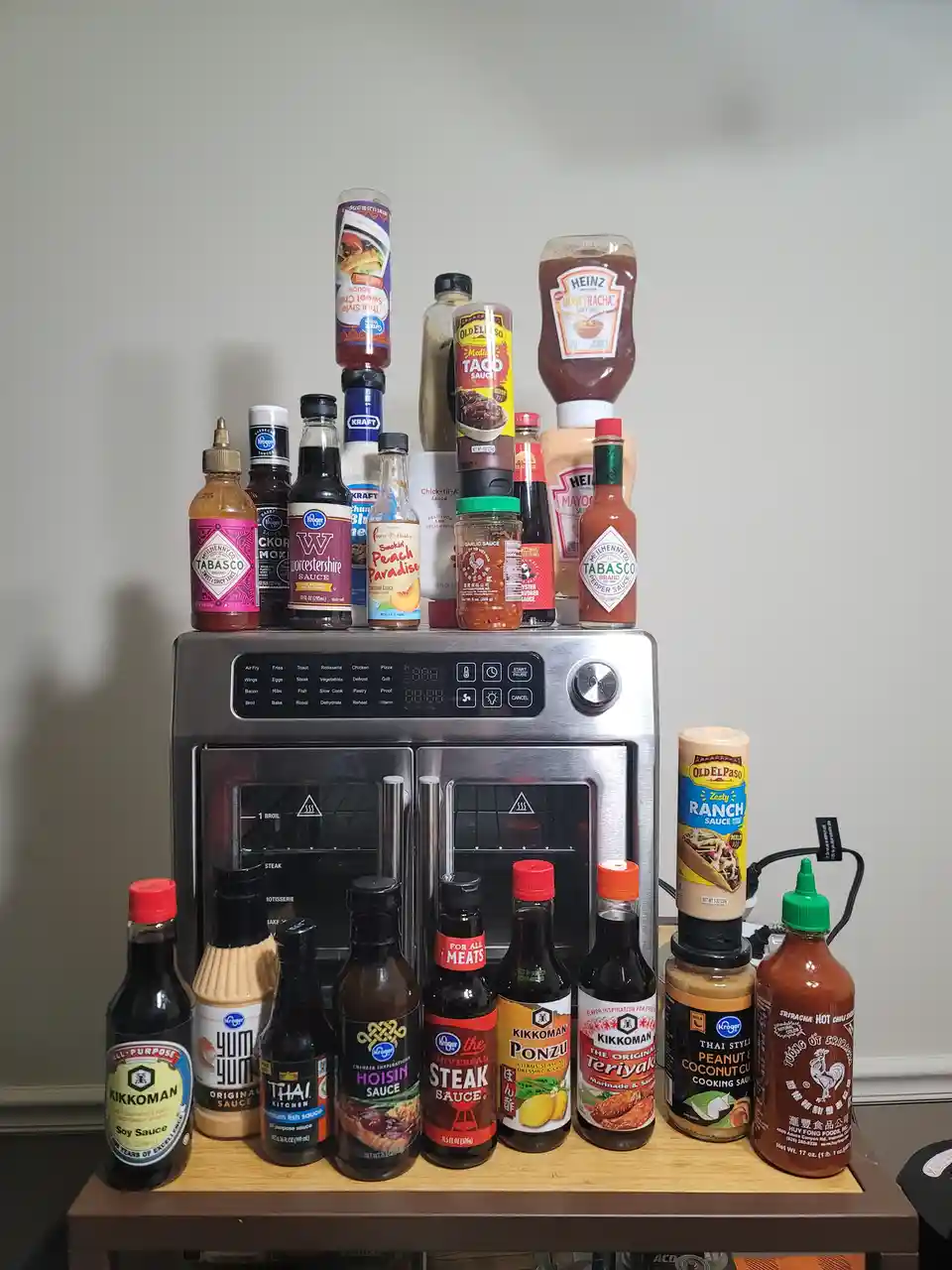 A stainless steel air fryer oven is surrounded by dozens of condiment bottles and sauce jars, including Tabasco, Kikkoman…