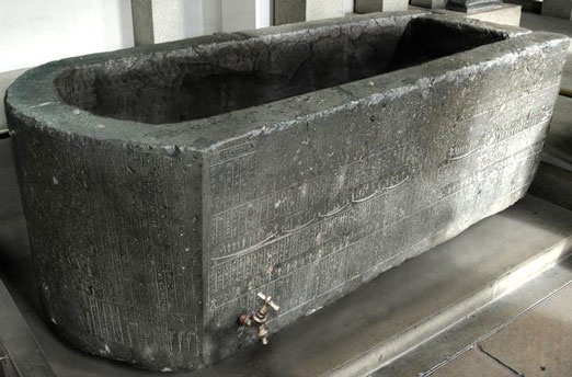 A large, ancient-looking stone bathtub with inscriptions carved into its side, placed on a modern metal platform. A small…