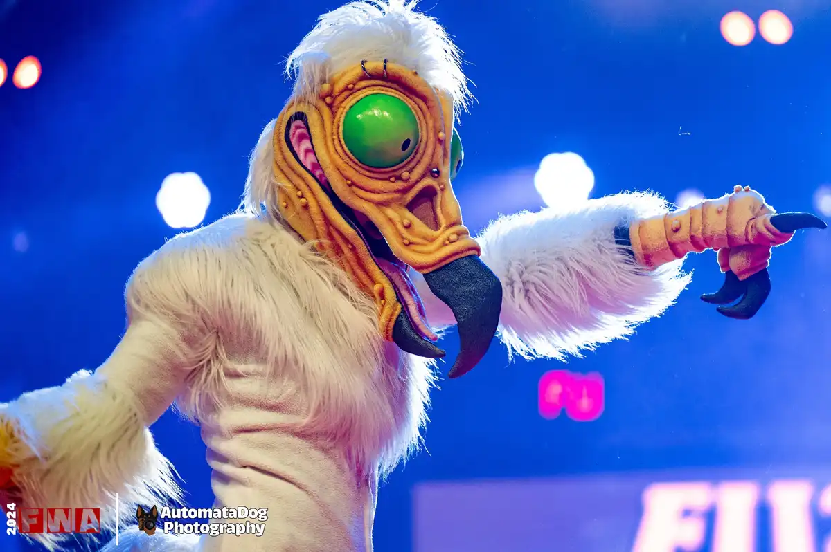 A performer in a furry, white costume with a large, orange, one-eyed alien mask and black beak-like mouthpiece points…