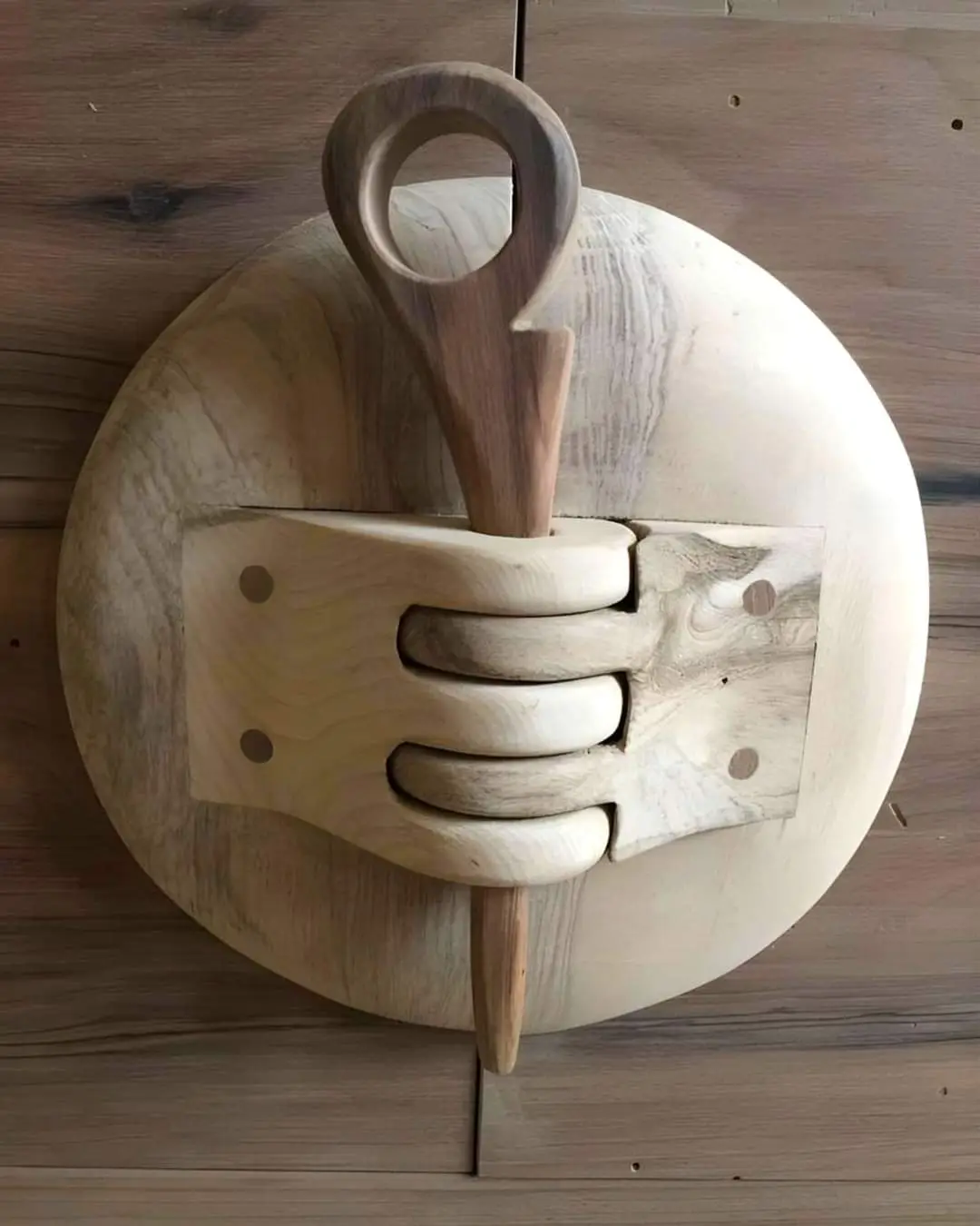 A wooden circular object with a carved hand gripping a wooden spoon handle, set against a wooden background. The overlaid…