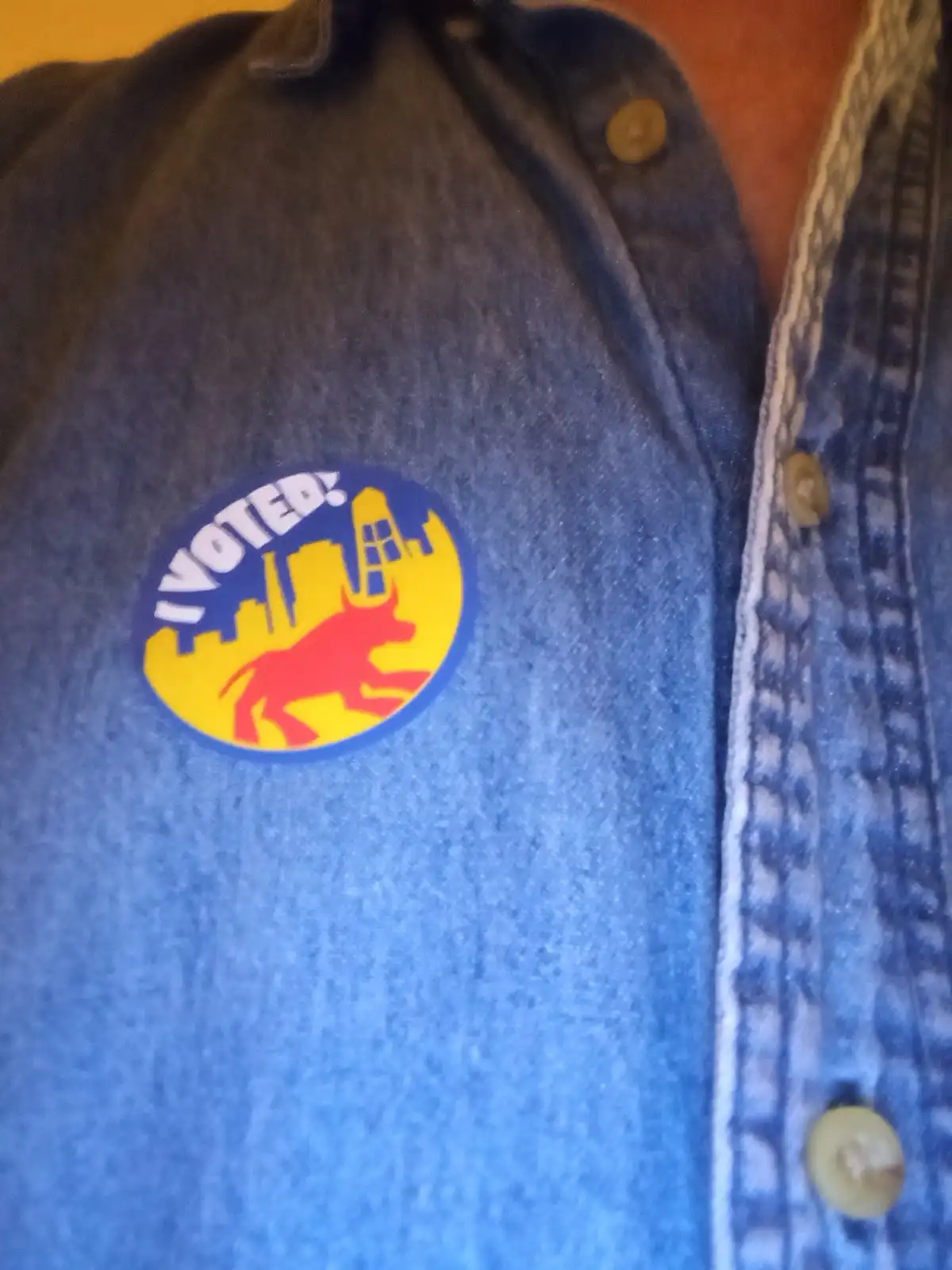 A close-up of a blue denim shirt with a circular 'I VOTED!' sticker on the chest. The sticker features a yellow background…