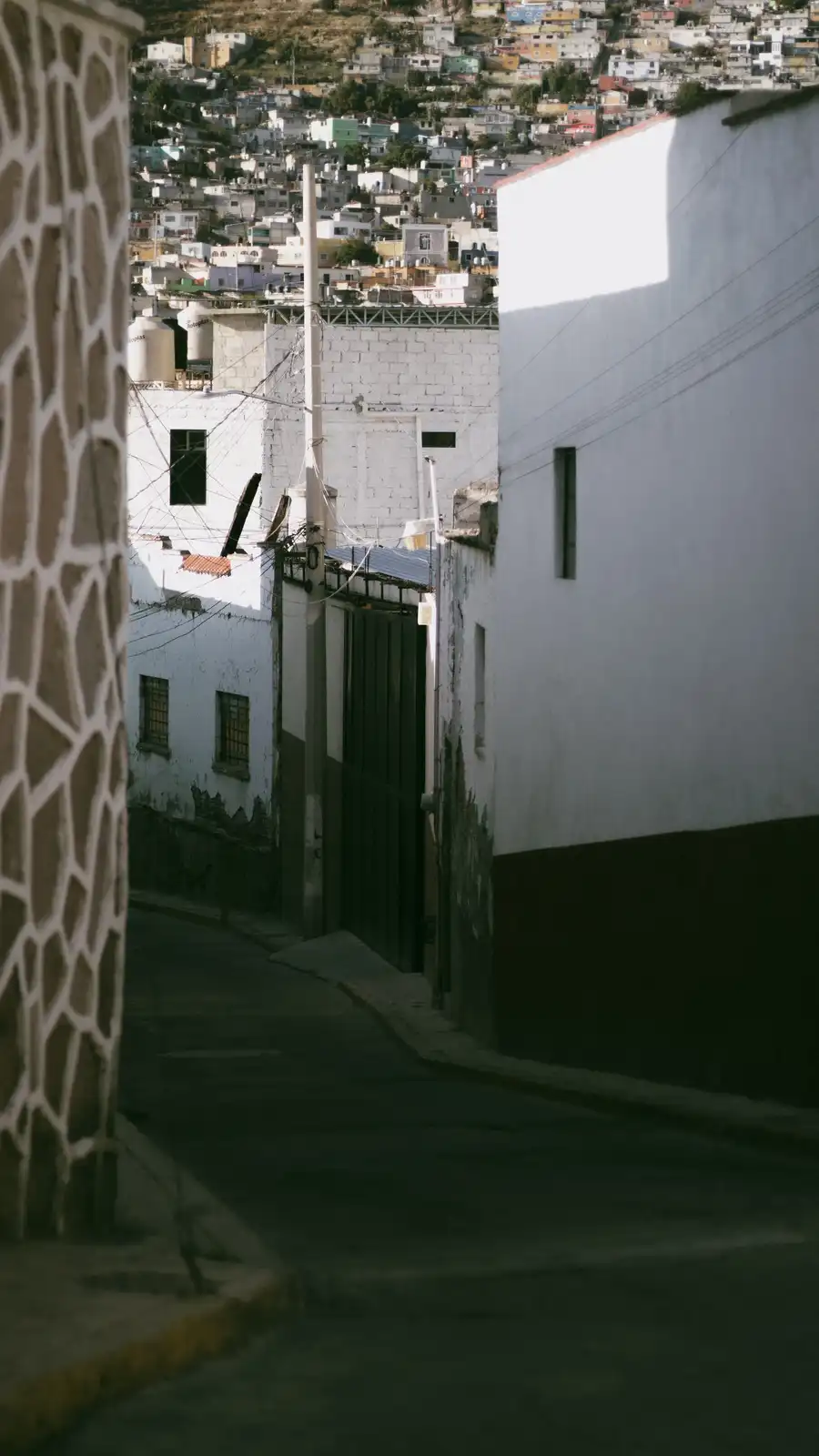 A narrow, shadowed alleyway framed by white and patterned walls, leading to a sunlit hillside of colorful buildings…