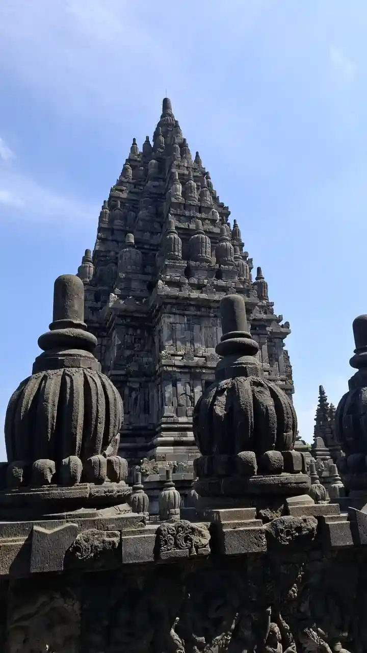 A low-angle photo shows the intricate, dark stone architecture of a historic temple, likely Prambanan in Indonesia, with…
