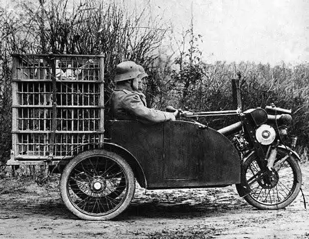 A black-and-white historical photo shows a soldier in a helmet driving a motorcycle with a sidecar. Attached to the back is…