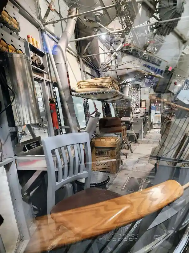 The image shows the interior of a vintage aircraft, with visible metal framework, fuel tanks, control panels, and a wooden…