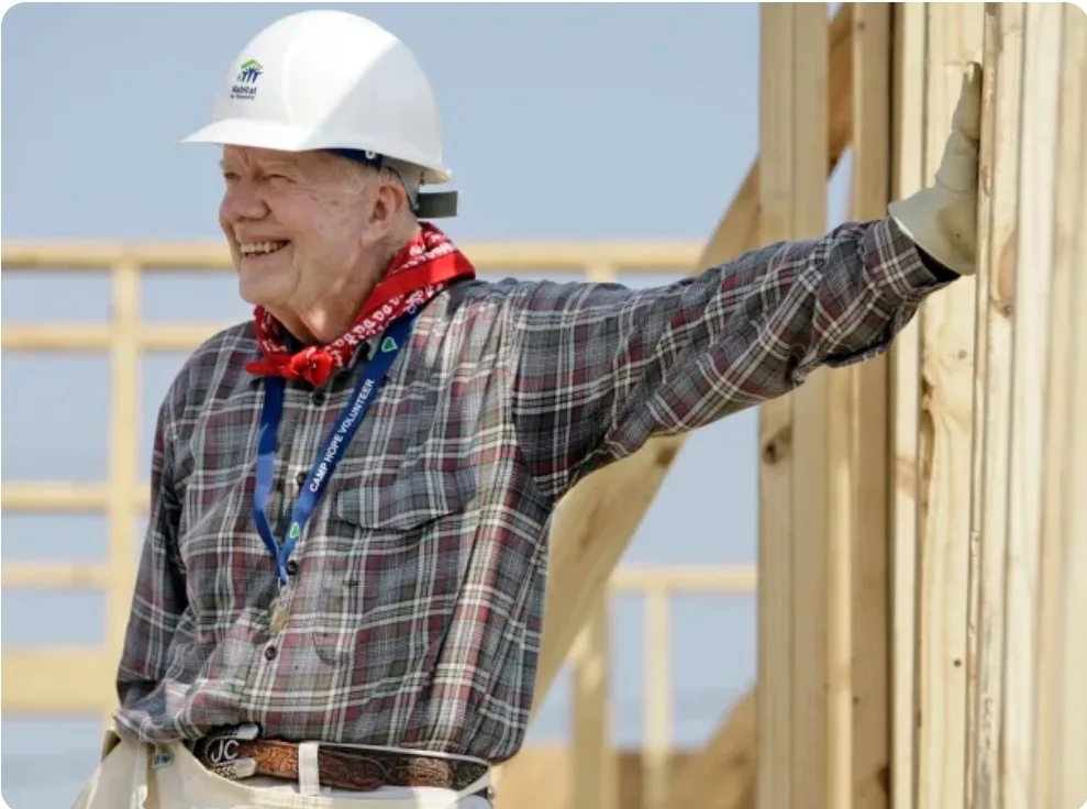 a smiling older man wearing a white hard hat with a 'Build' logo, a red bandana, a plaid shirt, and a 'Camp Hope Volunteer'…