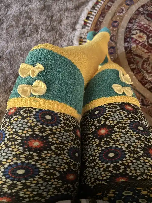 A close-up photo shows a person's feet wearing colorful, fuzzy socks with yellow bows on a teal cuff, paired with patterned…