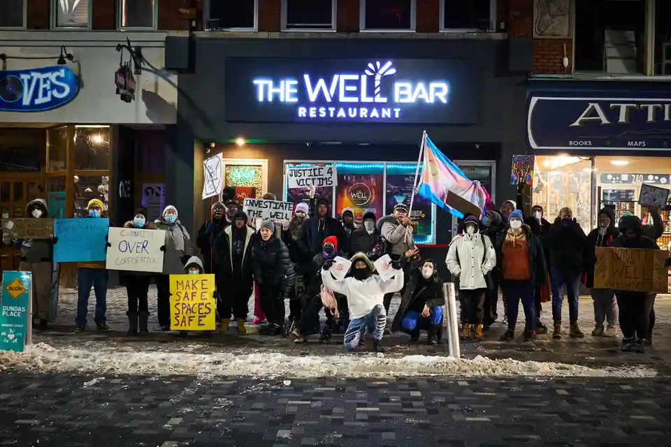 A nighttime photo shows a group of protesters gathered outside 'The Well Bar Restaurant,' holding signs demanding justice…