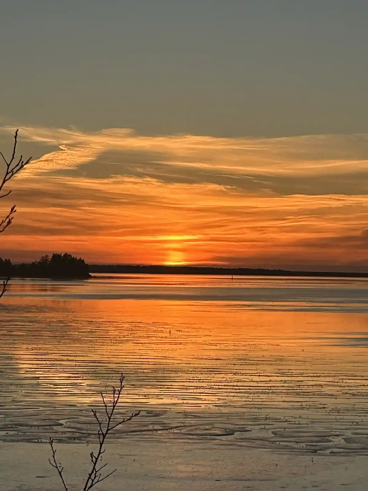 A serene sunset over a frozen lake with orange and yellow hues reflecting on the ice, framed by bare tree branches. The…