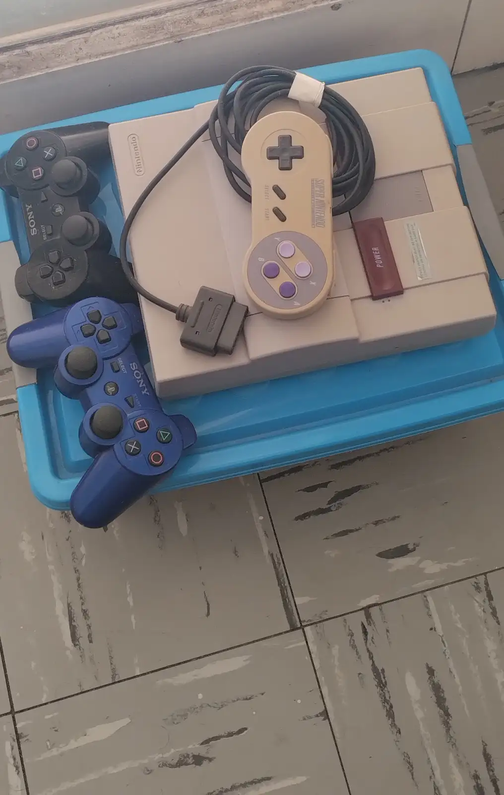 A beige Super Nintendo Entertainment System console with its controller and a black and blue Sony PlayStation controller are…