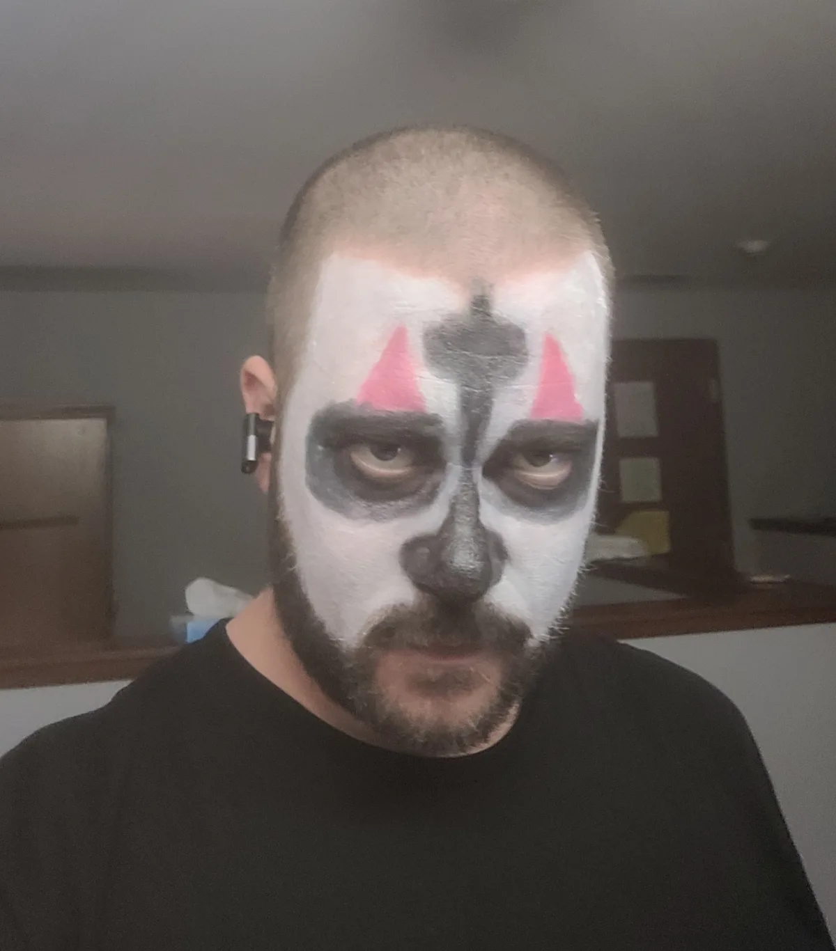 A man with a shaved head and beard wears dramatic white, black, and pink face paint resembling a clown or jester, with a…