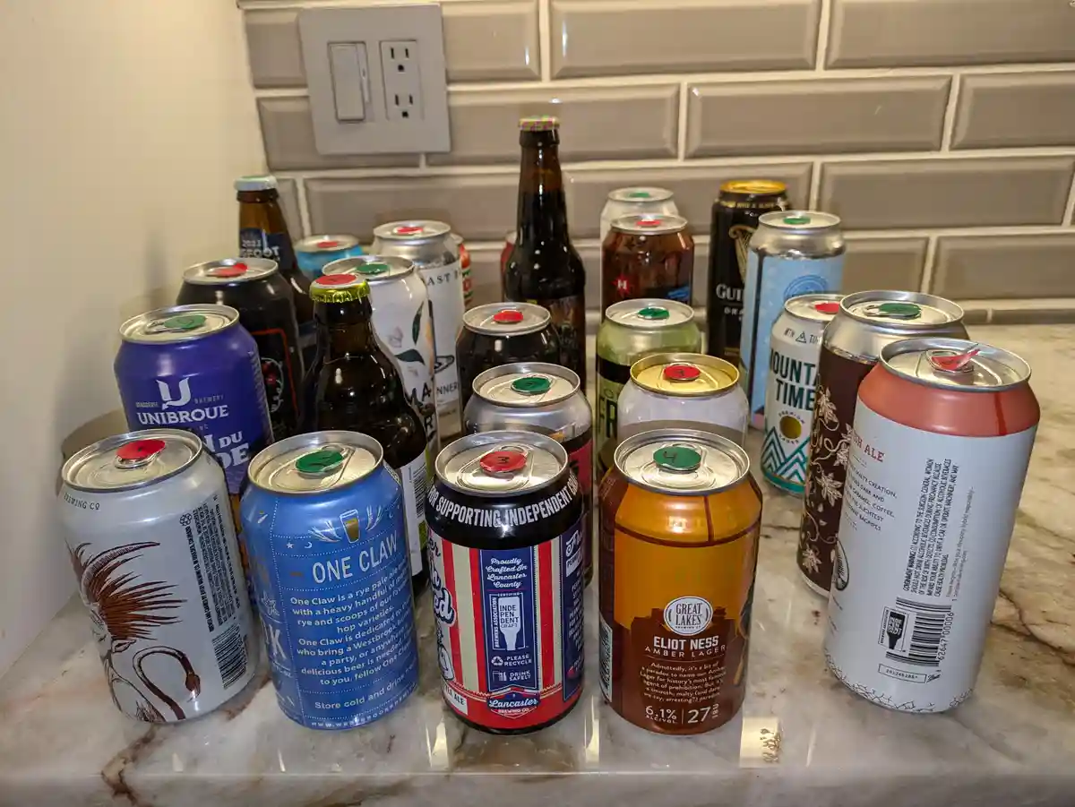 A countertop is filled with numerous beer cans and bottles, many with pull tabs marked with red or green numbers. The…