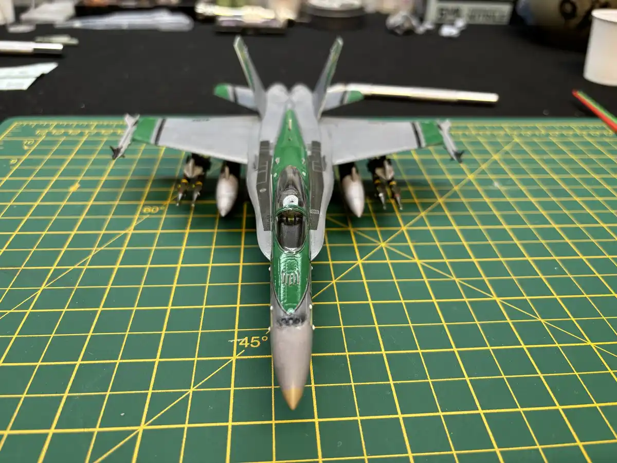 A detailed scale model of a fighter jet, painted green and grey with white missiles, sits centered on a green cutting mat…